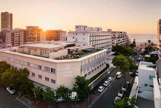 Hotellbygning ved sjøen omgitt av andre bygninger i solnedgang. (AI-generert tekst)