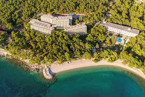 Flyfoto av hotellkompleks omgitt av skog og strand. (AI-generert tekst)