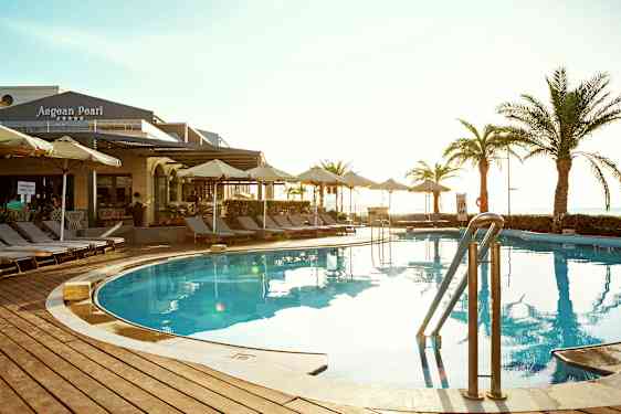 Utendørsbasseng med solsenger og parasoller rundt. Bygning med skilt 'Aegean Pearl' er synlig i bakgrunnen. (AI-generert tekst)