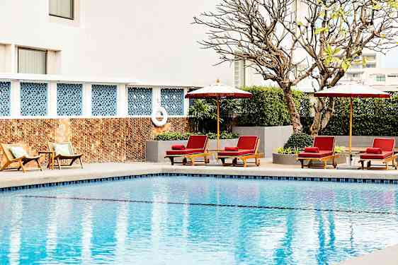 Et basseng med solsenger og parasoller ved kanten, omgitt av planter og en bygningsvegg i bakgrunnen. (AI-generert tekst)