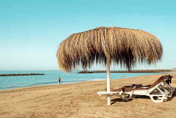 Bilde av en sandstrand med to solsenger under en halmparasoll og personer som går langs vannet. (AI-generert tekst)