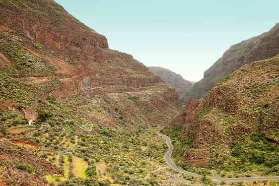 En smal vei slynger seg gjennom en dal omgitt av bratte fjell med grønn vegetasjon. (AI-generert tekst)