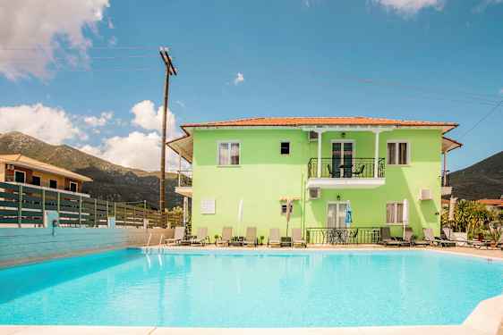 En lysegrønn bygning med balkonger og solsenger, foran et utendørs svømmebasseng. (AI-generert tekst)