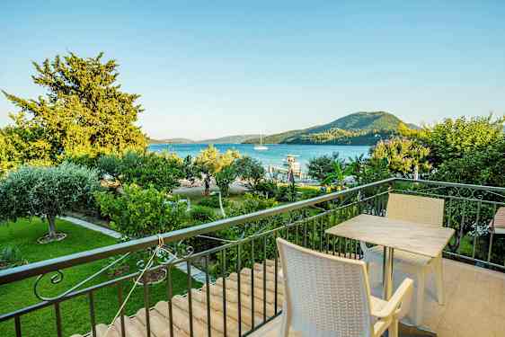 En balkong med bord og stoler, utsikt over en grønn hage, havet og fjell i bakgrunnen. (AI-generert tekst)