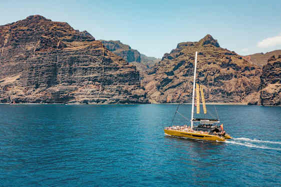 En katamaran seiler nær høye klipper, med klart blått vann i forgrunnen. (AI-generert tekst)