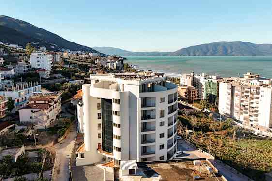 Et høyhus nær sjøen, omgitt av bygninger og grøntområder. (AI-generert tekst)