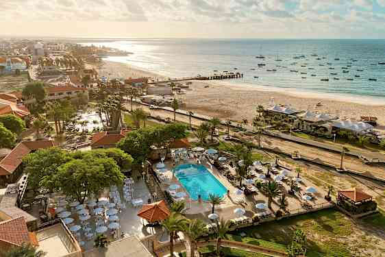 Luftfoto av en strand med solsenger og parasoller nær et basseng, omgitt av grøntområder. (AI-generert tekst)