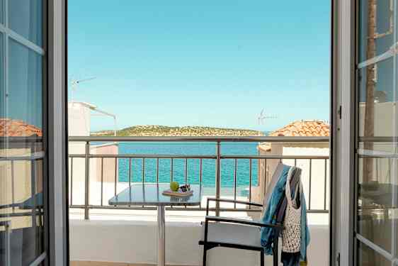 Utsikt fra et rom gjennom dører mot en balkong med bord, stol og klær, samt hav og landskap i bakgrunnen. (AI-generert tekst)
