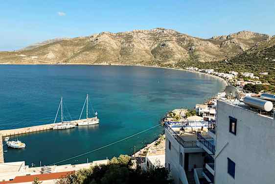 Utsikt over en kyst med små bygninger og fjell i bakgrunnen. Båter ligger fortøyd ved brygga i det klare vannet. (AI-generert tekst)