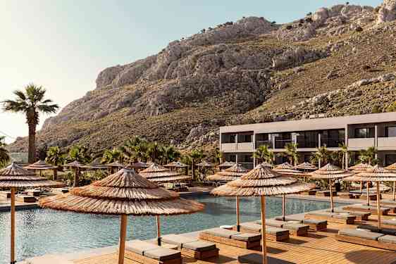 En moderne bygning med balkonger ligger ved siden av et svømmebasseng omgitt av solsenger og parasoller. I bakgrunnen er bergknauser og palmer. (AI-generert tekst)