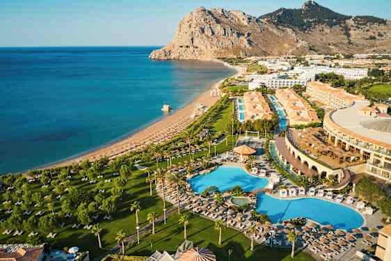 Luftbilde av et hotellområde med bassenger, grønne områder og en nærliggende strand ved sjøen. (AI-generert tekst)