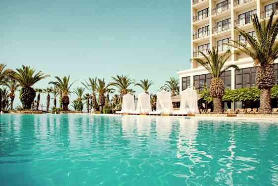 Utendørsbasseng med et hotell i bakgrunnen og omkringliggende palmer. (AI-generert tekst)