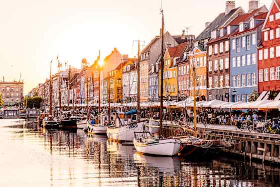 Fargerike bygninger langs en kanal med båter fortøyd i vannet og mennesker som sitter ved bord på kaien. (AI-generert tekst)