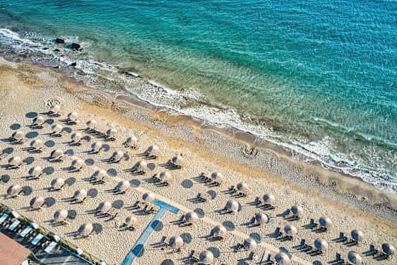 Luftfoto av en sandstrand med solsenger og parasoller, med kystlinjen og blått hav i bakgrunnen. (AI-generert tekst)