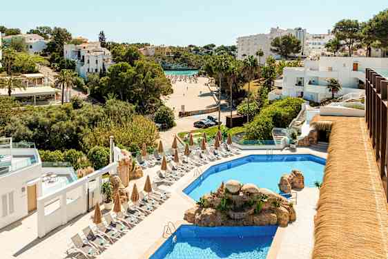 Hotellområde med et basseng omgitt av solsenger og parasoller, i bakgrunnen ses en strand og hvitmalte bygninger. (AI-generert tekst)