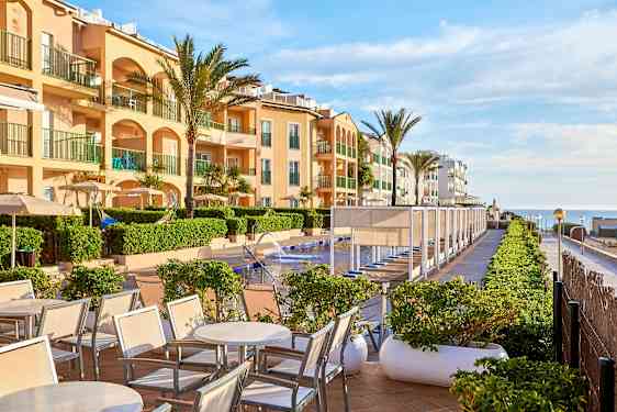 Et bilde av et hotellbasseng med solsenger og parasoller, omgitt av bygninger og palmer. (AI-generert tekst)