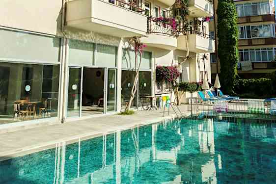 Et basseng foran en bygning med balkonger og store glassvinduer. Bildene viser også stoler og bord samt parasoller ved bassengområdet. (AI-generert tekst)