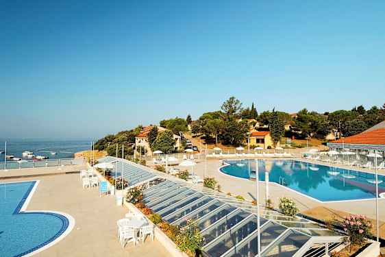 En utsikt over et utendørs svømmebasseng omgitt av en terrasse med solsenger og parasoller. I bakgrunnen er sjøen samt bygninger og vegetasjon. (AI-generert tekst)