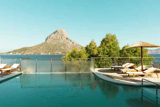 Et utendørsbasseng med solsenger og parasoll, plassert nær havet med en ås i bakgrunnen. (AI-generert tekst)