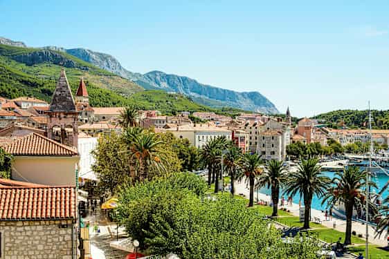 Bildet viser en pittoresk middelhavsby med rødteglstak, kirketårn, palmer og grønne fjell i bakgrunnen. En småbåthavn vises også ved vannet. (AI-generert tekst)