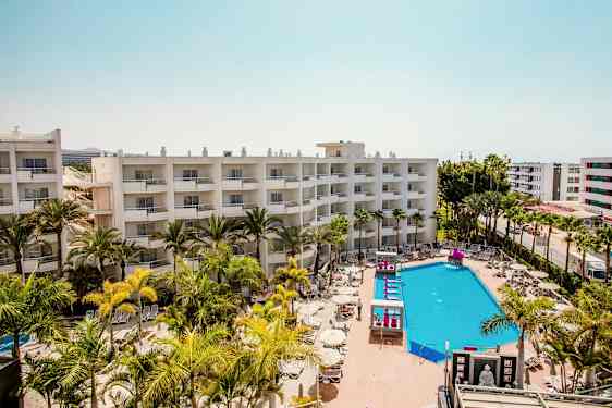 Hotellbygninger med balkonger, et bassengområde med solsenger og parasoller omgitt av palmer. (AI-generert tekst)