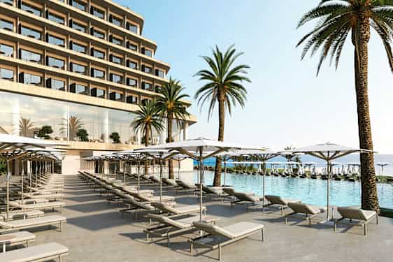 Bildet viser et hotellbasseng med solsenger og parasoller i forgrunnen, samt store palmer og hotellfasaden i bakgrunnen. (AI-generert tekst)