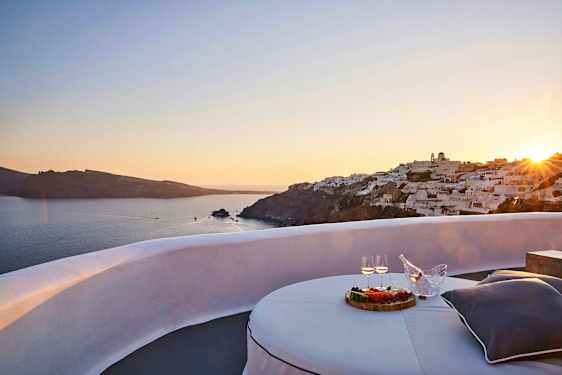 Utsikt over en solnedgang ved sjøen med hvite bygninger på en klippe og en terrasse med bord, mat og drikke i forgrunnen. (AI-generert tekst)