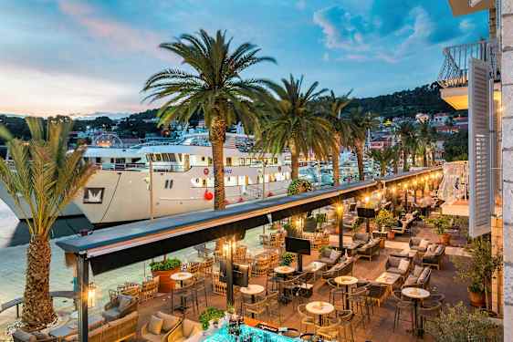 En utsikt over en utendørsrestaurant ved en havn med palmer og store båter. (AI-generert tekst)