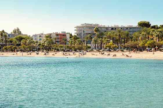 En strandlinje med klart blått vann og solbadende mennesker. I bakgrunnen er det palmer og bygninger. (AI-generert tekst)