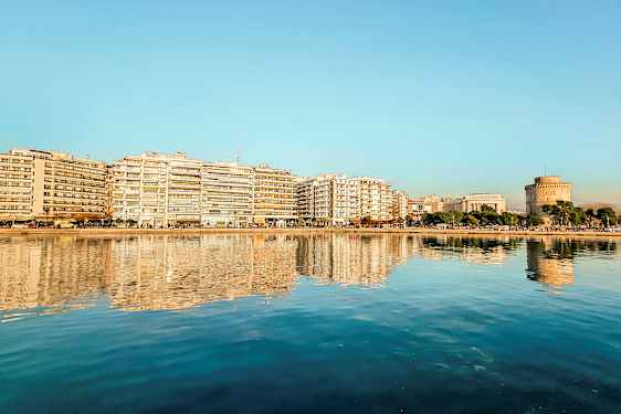 En by ved vannet med bygninger og et rundt tårn reflektert i det rolige vannet. (AI-generert tekst)