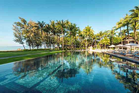 Et utendørsbasseng omgitt av grøntområder og palmer, med utsikt over stranden og havet. (AI-generert tekst)