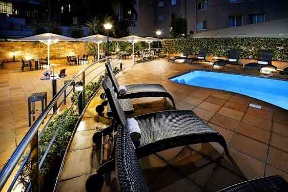 Hotellens bassengområde med solsenger og parasoller ved et opplyst flisebelagt uteområde. (AI-generert tekst)