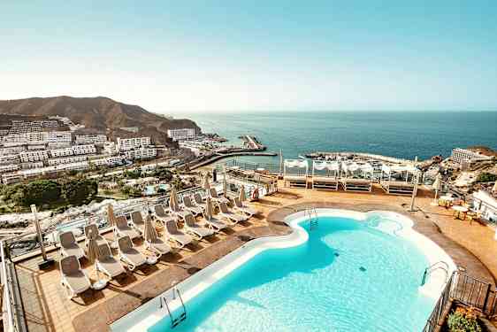 En terrasse med et basseng og solsenger, med utsikt over havet og en kystby. (AI-generert tekst)