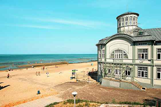 En strand med solbadende mennesker ved en rolig kystlinje, samt en større bygning med klassisk arkitektur ved stranden. (AI-generert tekst)