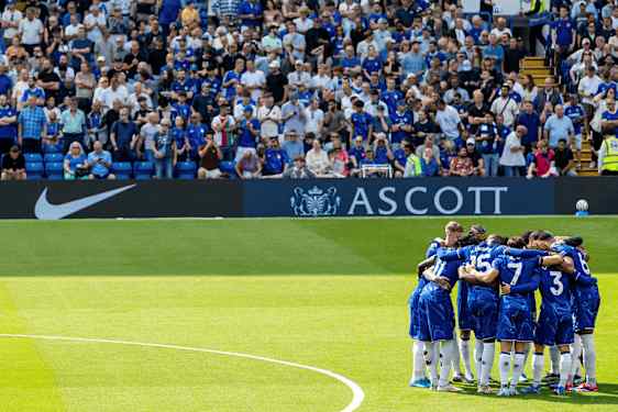 Spillere i blå fotballdrakter står samlet i en sirkel på grønt gress med publikum i bakgrunnen. (AI-generert tekst)