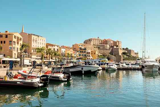 En småbåthavn med båter fortøyd ved kaien, omgitt av fargerike bygninger og en historisk borg på en høyde i bakgrunnen. (AI-generert tekst)