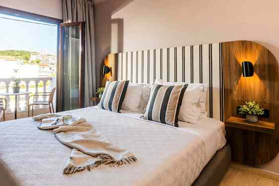 Et hotellrom med en dobbeltseng, stripete pynteputer og et bord med stoler ved en åpen balkongdør. (AI-generert tekst)