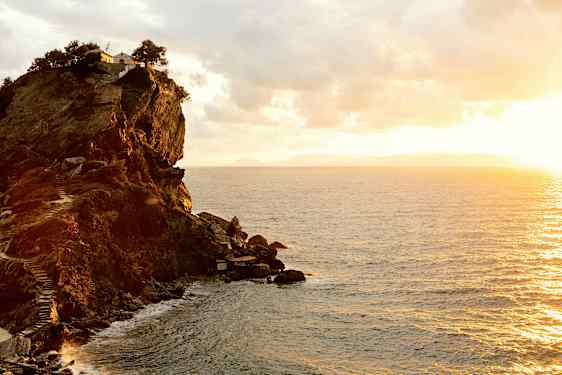 En klippe ved kysten med en bygning og trær på toppen. Havet sees under en solnedgang. (AI-generert tekst)