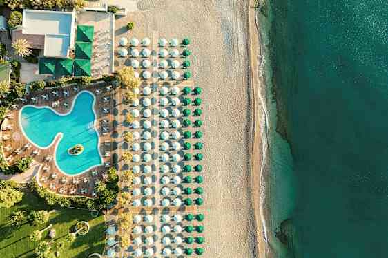 Flyfoto av hotellbasseng med omkringliggende solsenger, ved siden av en strand med parasoller og hav. (AI-generert tekst)