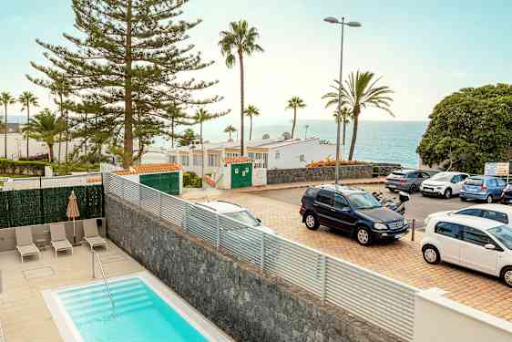 Et bilde av et utendørsmiljø med et basseng i forgrunnen, parkerte biler, trær, bygninger og utsikt over havet i bakgrunnen. (AI-generert tekst)