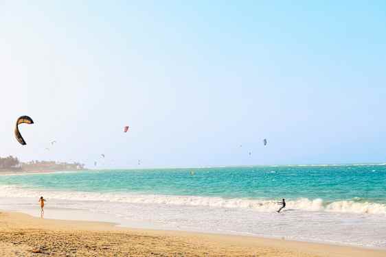 En strand med turkist hav og mange kitesurfere. (AI-generert tekst)