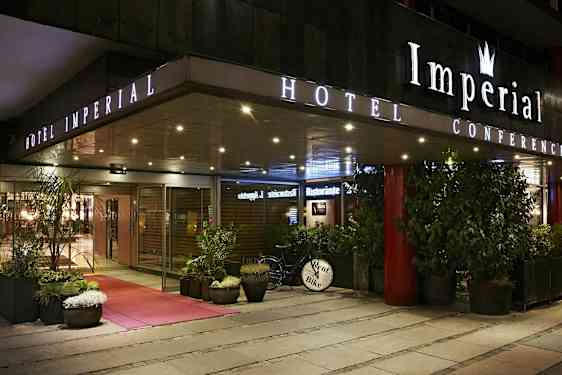 Utvendig bilde av et hotell om kvelden med skilt som viser 'Hotel Imperial', omgitt av planter og en sykkel med skiltet 'Rent Bike'. (AI-generert tekst)