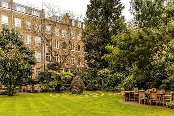 En grønn hage med bord og stoler og et steinmonument i midten, omgitt av bygninger. (AI-generert tekst)