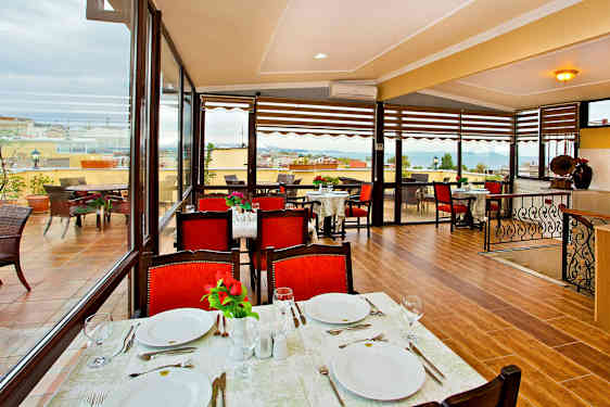 En restaurant med bord dekket for måltid, røde stoler og utsikt over en terrasse samt havet. (AI-generert tekst)