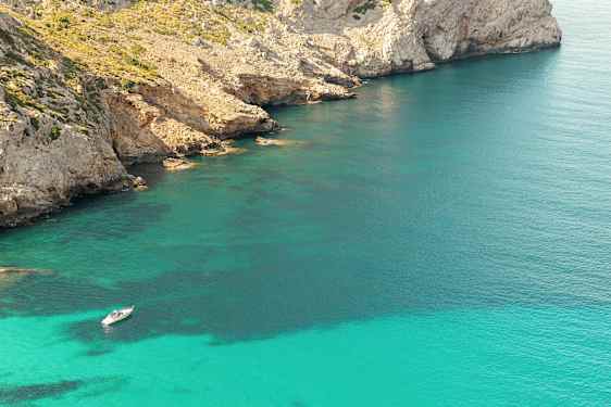 En båt ligger ankrert i klart turkis vann nær en steinete kystlinje med klipper. (AI-generert tekst)