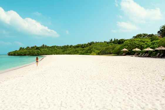 En kvinne går på en sandstrand med turkis vann og grønn vegetasjon i bakgrunnen. (AI-generert tekst)