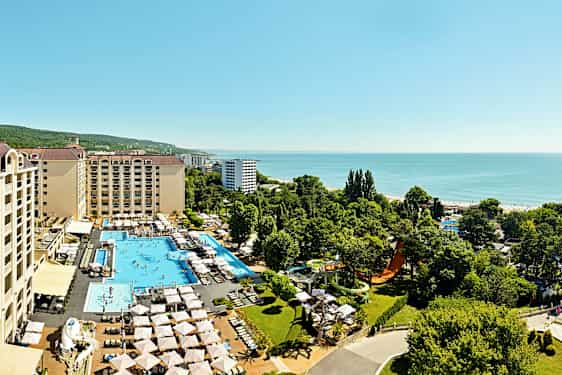 Luftfoto av hotellkompleks med bassenger, solsenger og grøntområder, plassert nær sjøen. (AI-generert tekst)