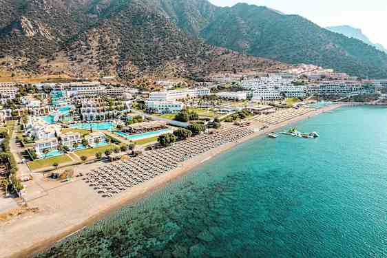 Flyfoto av en kyst med hotellkompleks ved stranden, bassenger og solsenger. (AI-generert tekst)