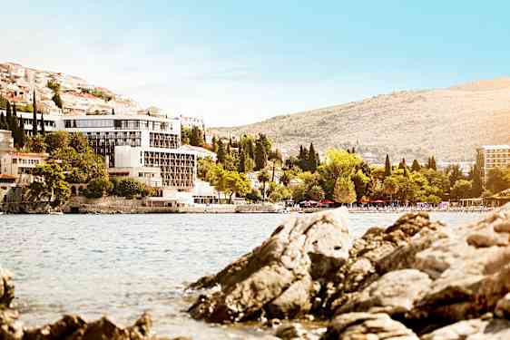 Utsikt mot en bygning ved en kystlinje med steinete strand og grønne trær. (AI-generert tekst)