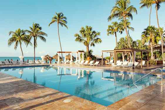 Et basseng omgitt av solsenger, palmer og en pergola, plassert nær sjøen med en klar blå himmel. (AI-generert tekst)